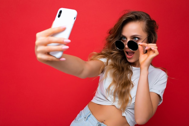 Primer plano de una mujer joven hermosa increíble feliz que sostiene el teléfono móvil que toma la foto del selfie usando