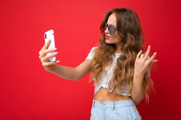 Primer plano de una mujer joven hermosa increíble feliz que sostiene el teléfono móvil que toma la foto del selfie usando