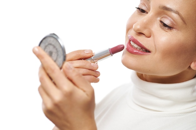 Primer plano de una mujer joven con hermosa sonrisa aplicando un lápiz labial en los labios y mirándose a sí misma en un pequeño espejo cosmético
