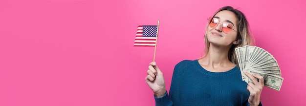 El primer plano de una mujer joven y hermosa con un suéter azul en gafas con dinero en dólares americanos y una bandera en la mano sobre un fondo rosa