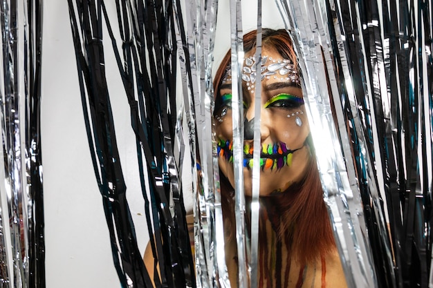 Primer plano de una mujer joven con maquillaje para halloween escondido entre tiras negras y plateadas brillantes