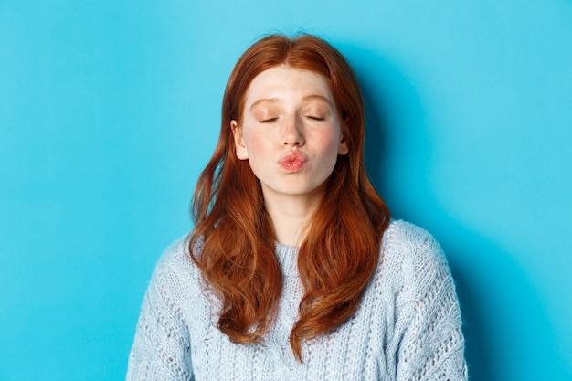 Primer plano de una mujer joven con el pelo ondulado rojo de pie con los ojos cerrados y los labios fruncidos, esperando un beso contra el fondo azul.