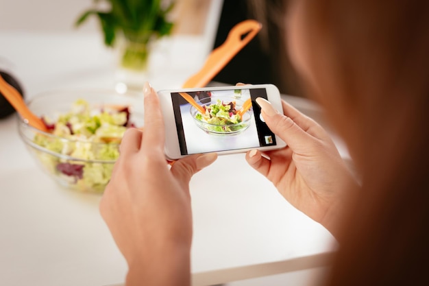 Primer plano de una mujer joven que toma una foto de una ensalada saludable con un teléfono inteligente para su blog. Vista trasera.