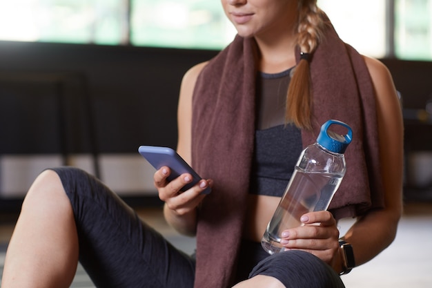 Primer plano de una mujer joven sentada con una botella de agua y usando su teléfono móvil después del entrenamiento deportivo