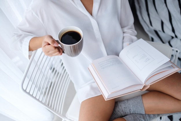 Primer plano de una mujer leyendo un libro y bebiendo café caliente en el dormitorio en casa por la mañana