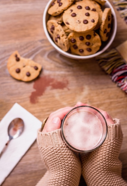 Primer plano de una mujer manos sosteniendo un vaso de bebida caliente sobre una mesa con galletas con chispas de chocolate en un tazón de estrellas