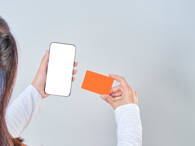 Foto primer plano de mujer mantenga smartphone, pantalla en blanco y tarjeta bancaria de crédito sobre fondo gris