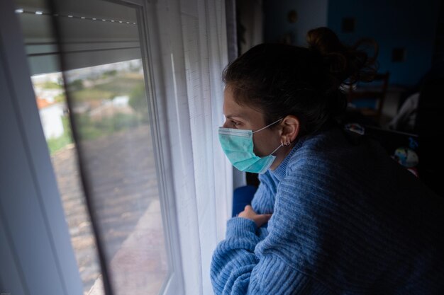 Foto primer plano de una mujer con máscara mirando por la ventana de su casa