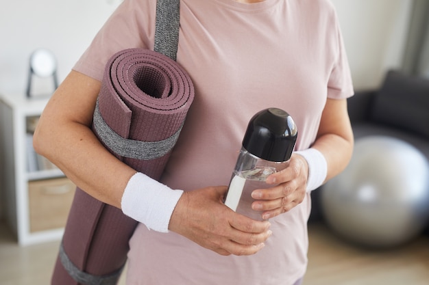 Primer plano de mujer mayor sosteniendo una colchoneta de ejercicios y una botella de agua que está lista para el entrenamiento deportivo