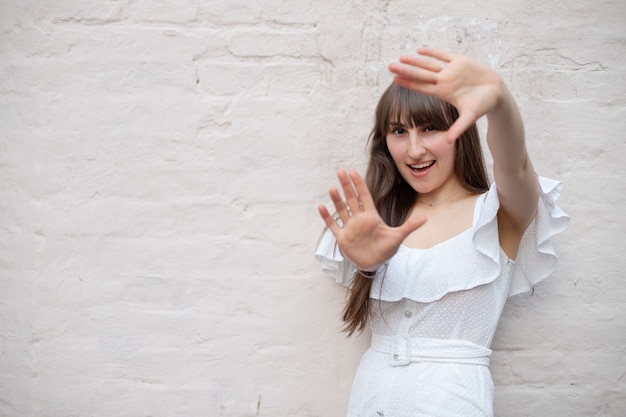 Primer plano de una mujer morena con el pelo largo con un vestido blanco hace un marco de sus manos y sonríe sobre un fondo de ladrillo monocromo. el lugar para tu diseño