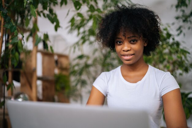 Primer plano de una mujer de negocios afroamericana positiva que trabaja en una computadora portátil sentada en el escritorio en la sala de la oficina en casa con diseño de biofilia