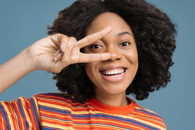 Primer plano de una mujer nigeriana sonriente que muestra el signo de la victoria mirando a la cámara aislada en el fondo azul