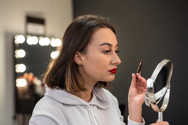 El primer plano de una mujer pinta su ceja con pintura con un hueso en un pequeño espejo de maquillaje, la niña se tiñe las cejas ella misma