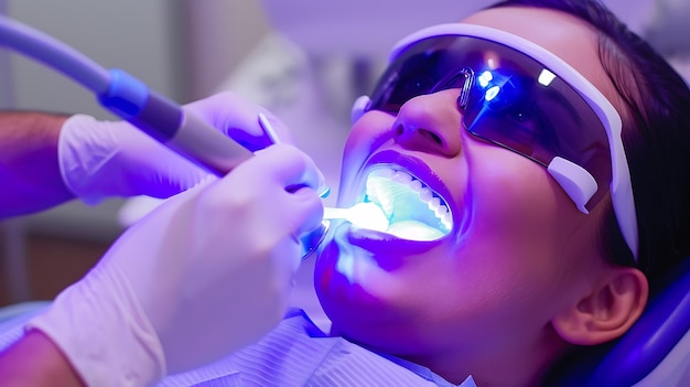 Un primer plano de una mujer que tiene sus dientes blanqueados con una luz azul Ella lleva gafas de protección y el dentista tiene una herramienta en la mano