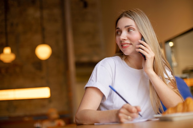 Primer plano de una mujer rubia satisfecha con camiseta blanca mira hacia un lado y habla por teléfono, mientras trabaja en el café contra un fondo de pared de ladrillo. lugar para tu diseño