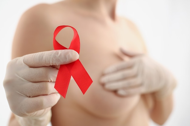 Primer plano de mujer sosteniendo una cinta roja, lucha contra el cáncer de mama, apoya a la mujer en la lucha contra la enfermedad. Mujer en topless sobre fondo borroso. Cáncer de mama, enfermedad, medicina, concepto de diagnóstico