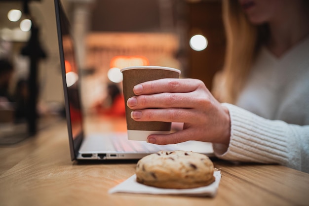 Primer plano de una mujer sosteniendo una taza de café mientras usa su computadora portátil en una cafetería