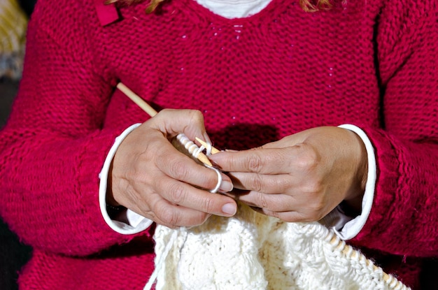 Primer plano de mujer en suéter rojo tejiendo hilo de lana blanca con agujas a mano.