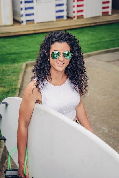 Primer plano de una mujer surfista morena rizada con top blanco y gafas de sol caminando con una tabla de surf