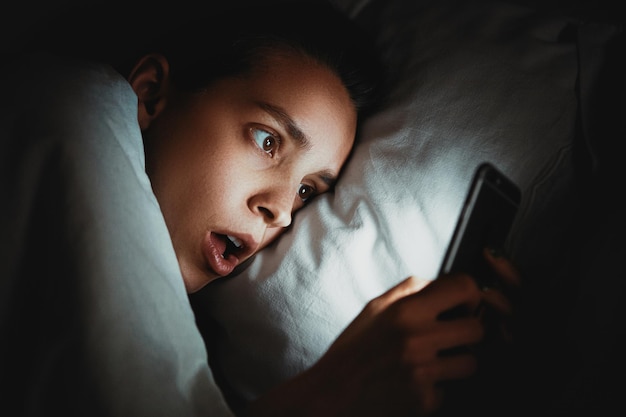 Primer plano de una mujer usando el teléfono mientras está acostada en la cama en un cuarto oscuro
