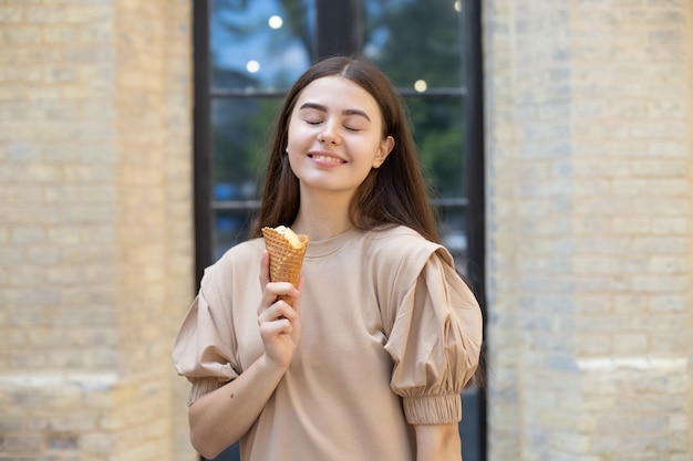 Primer plano de una niña morena sonriente con los ojos cerrados comiendo helado en una taza de waffle contra un fondo de pared de ladrillo