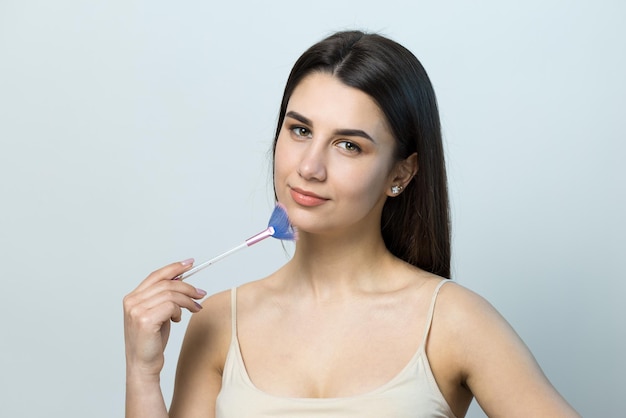 Primer plano de una niña en una parte superior clara sobre un fondo blanco haciendo un maquillaje facial Una mujer bonita sostiene un cepillo cosmético cerca de su cara y sonríe