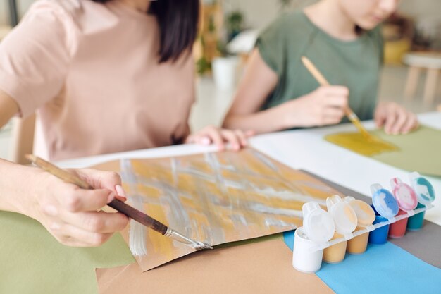 Primer plano de niñas irreconocibles sentadas en la mesa y pintar cuadros de pared con gouache