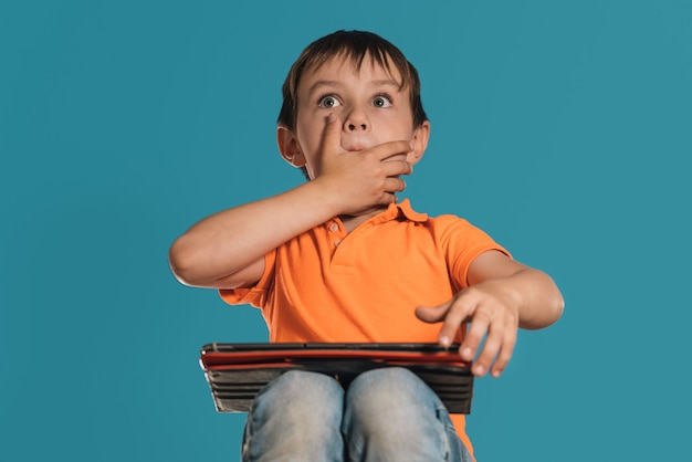 Foto primer plano de un niño con una camiseta naranja con una tableta sobre un fondo azul expresión de sorpresa