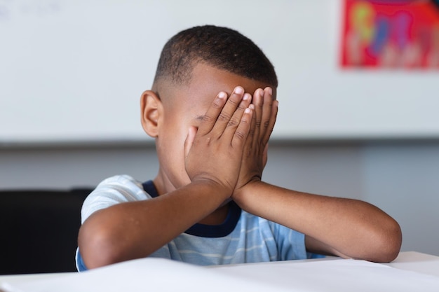 Primer plano de un niño de escuela primaria afroamericano con las manos cubriendo la cara sentado en el escritorio en clase