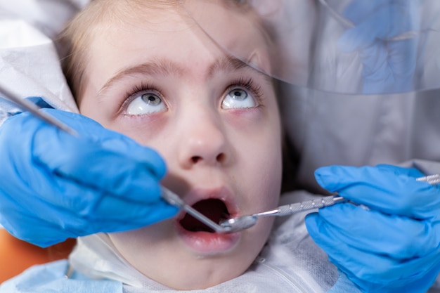 Primer plano de un niño que recibe tratamiento de caries del dentista durante el brote de covid19. Dentista en traje de coronavirus con espejo curvo durante el examen de los dientes del niño.