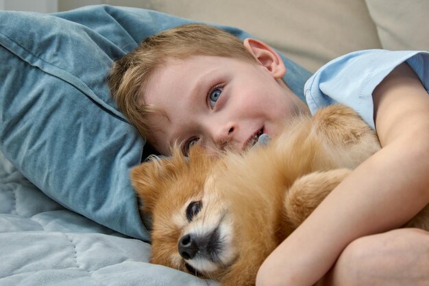 Primer plano de un niño rubio sonriente abrazando suavemente a un lindo perro rojo Un niño feliz con su Spitz alemán está acostado en una cama con almohadas Amistad con una mascota encantadora