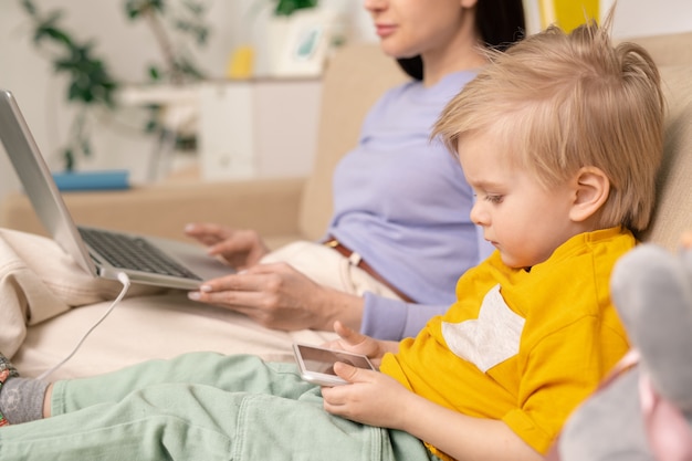 Foto primer plano de un niño serio concentrado sentado en el sofá y usando un gadget mientras la madre trabaja con la computadora portátil