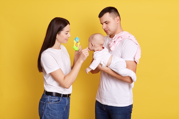 Primer plano de un padre acariciando al bebé en las manos del padre y la mamá de pie junto a ellos dando chupete para el bebé y mostrando el juguete al recién nacido, familia con ropa casual y posar contra la pared amarilla.