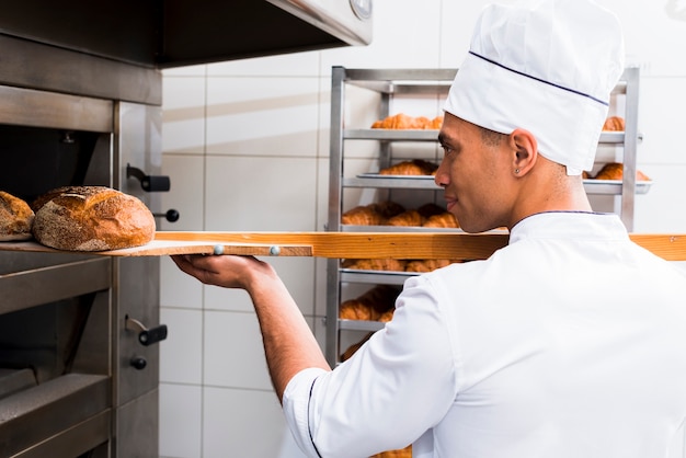 Primer plano de panadero de sexo masculino en uniforme sacando con pala pan recién horneado del horno