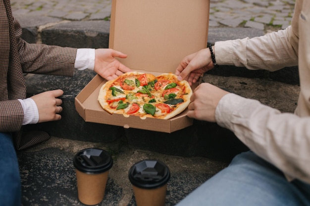 Un primer plano de una pareja agarrando rebanadas de pizza de una caja en el exterior, un hombre y una mujer con las manos tomando pizza.