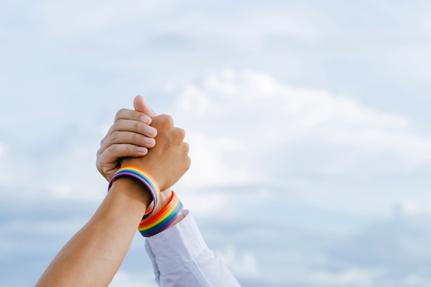 Primer plano de una pareja gay cogidos de la mano con una pulsera de arco iris en el cielo