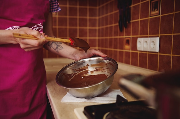 Primer plano de un pastelero irreconocible en delantal rosa mezclando masa de chocolate derretido en un tazón con una espátula de madera de pie en la cocina de su casa