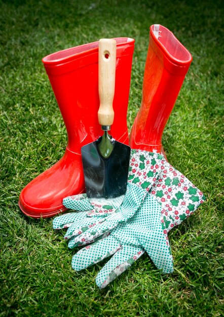 Primer plano de una pequeña pala, guantes y botas de goma roja recostada sobre la hierba verde