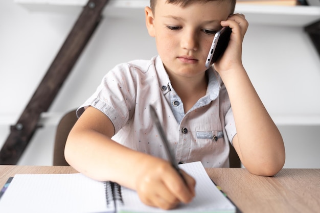 Primer plano de un pequeño estudiante hablando por un teléfono celular Un niño se sienta en un escritorio habla en un teléfono inteligente y toma notas en un cuaderno Concepto de tecnología y educación