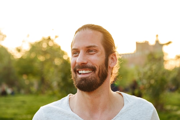 Primer plano de perfil de hombre feliz alegre de 30 años con el pelo atado en camiseta blanca riendo con una hermosa sonrisa, mientras descansa en el parque verde