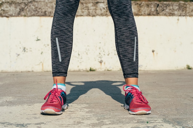 Primer plano de las piernas femeninas atléticas en zapatillas de deporte rojas en un cojín concreto al aire libre en luz del sol