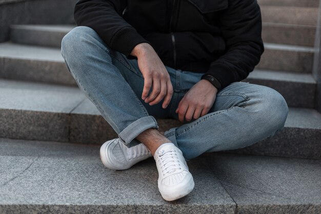 Primer plano de las piernas masculinas. Hombre con una elegante chaqueta negra en elegantes jeans azules en zapatillas de cuero blanco de moda está descansando sentado en una escalera de piedra vintage en la ciudad