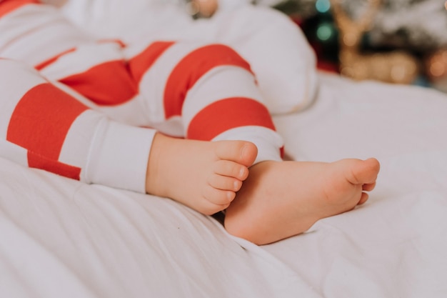 Primer plano de los pies descalzos de un niño en pijama rojo y blanco acostado en la cama sobre una sábana blanca. tacones navideños. foto de alta calidad