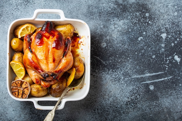 Primer plano de pollo orgánico crujiente jugoso entero horneado o asado con papas, ajo, limón en una cazuela blanca sobre fondo rústico de hormigón, vista superior, espacio para texto