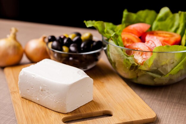 Foto primer plano de queso feta en tablero de madera sobre una mesa con ensalada de verano, cebollas y recipiente de vidrio lleno de aceitunas