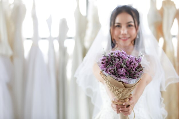 Primer plano de ramo de flores de boda púrpura celebrado en manos de la bella novia asiática en vestido blanco con velo de pelo transparente permanente mirada sonriente a cámara en fondo borroso en vestidor.