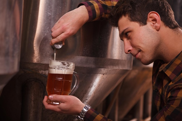 Primer plano recortado de un cervecero que vierte deliciosa cerveza artesanal fresca en una taza del tanque de cerveza en la cervecería