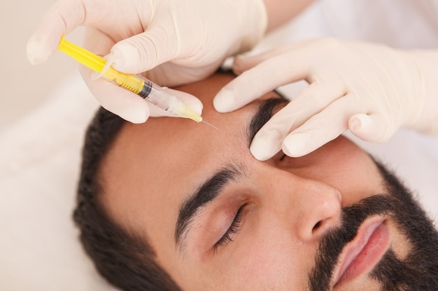 Primer plano recortado de una esteticista inyectando relleno facial en las arrugas de la frente del cliente masculino
