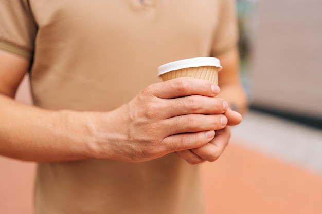 Primer plano recortado de un joven irreconocible con camiseta sosteniendo en las manos una taza con café para llevar para ir de pie posando con en la calle de la ciudad