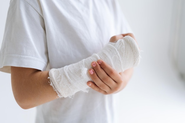 Primer plano recortado de una niña irreconocible con la mano rota envuelta en una venda de yeso masajeando suavemente la muñeca lesionada sobre fondo blanco aislado